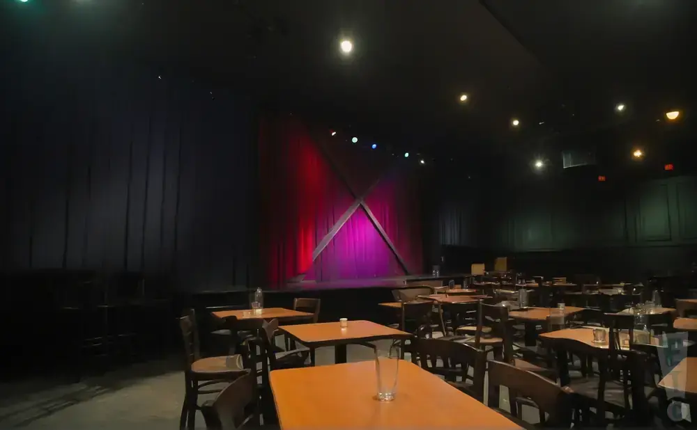 A comedy club stage with red curtains and tables with chairs in the foreground.