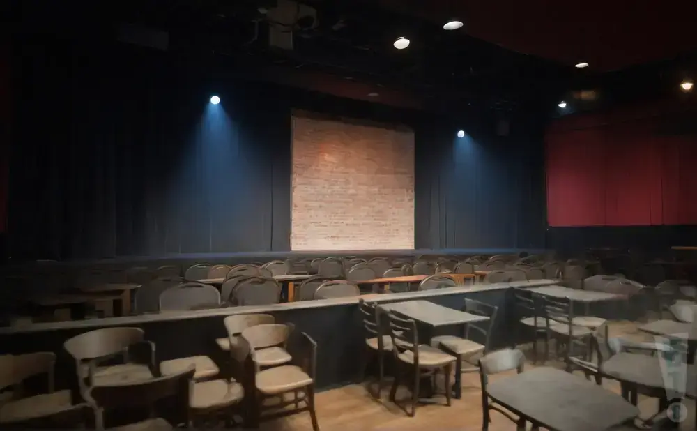 An empty theater with stage, brick backdrop, and rows of chairs and tables.