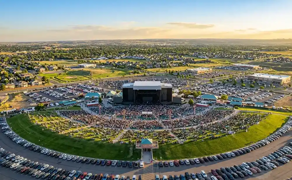 An aerial view of an outdoor concert venue with a large crowd, stage, and surrounding parking lots at sunset.