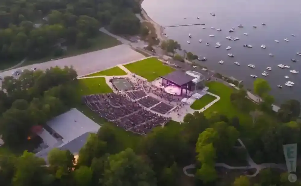 Aerial view of an outdoor amphitheater with a stage and audience, located by a body of water with many boats.