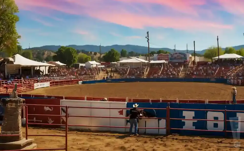 Rodeo arena with spectators, cowboys, and a bull under a colorful sunset sky.