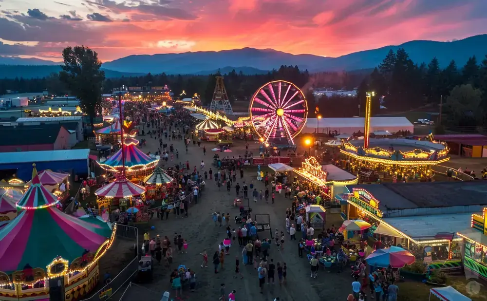 Aerial view of a bustling fair at sunset, with colorful rides and a crowd of people under a vibrant sky.