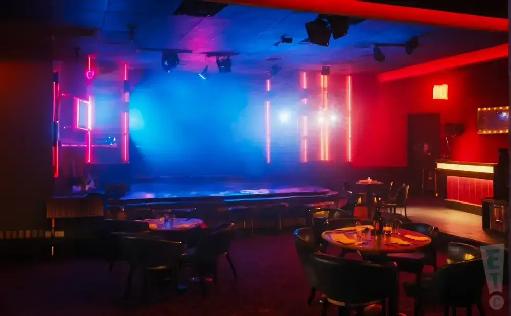 A nightclub with a stage, tables, chairs, and a bar, lit by red and blue neon lights.