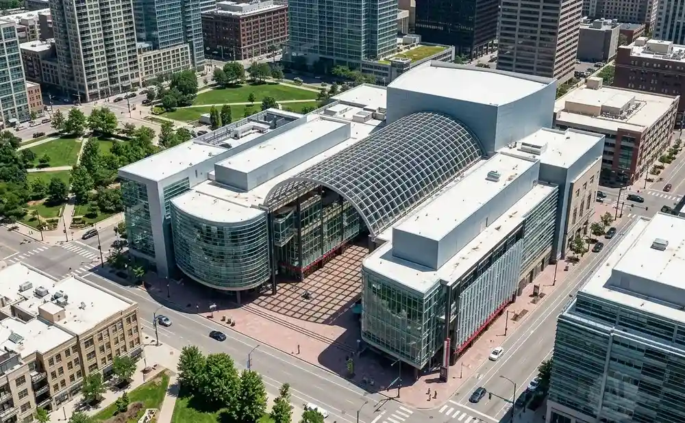 Aerial view of a modern convention center with a glass-arched roof, surrounded by city buildings and green spaces.