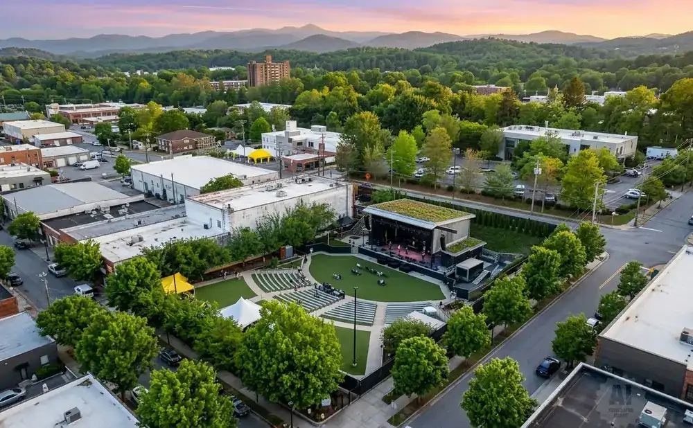Aerial view of an outdoor amphitheater with tiered seating and a stage, surrounded by trees and buildings at sunset.