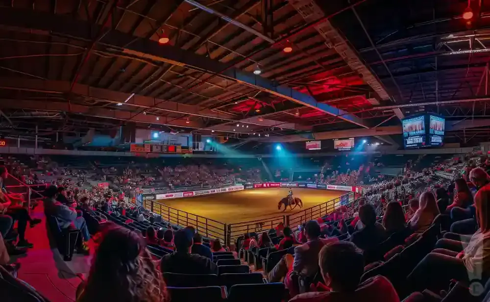 A rodeo event with a cowboy on horseback in a dirt arena, surrounded by stadium seating filled with spectators.