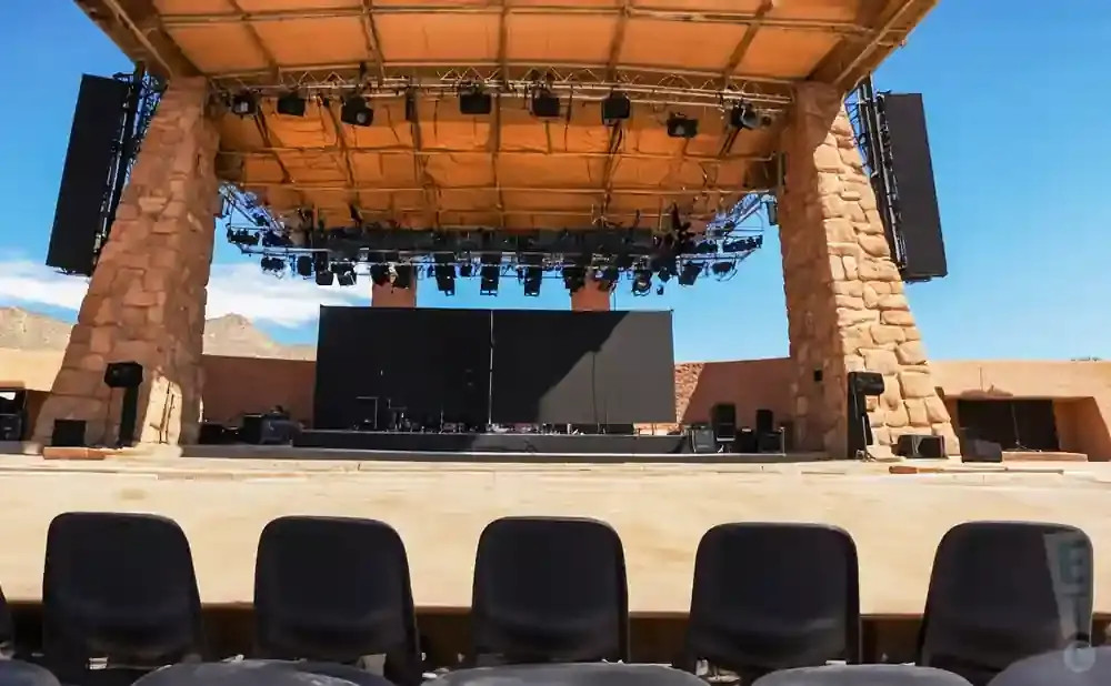 An outdoor amphitheater stage with a large screen, speakers, and lights.