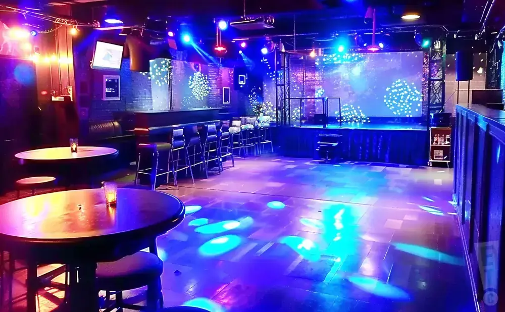A dimly lit club with a stage, bar, tables, and colorful lights projected onto the floor.