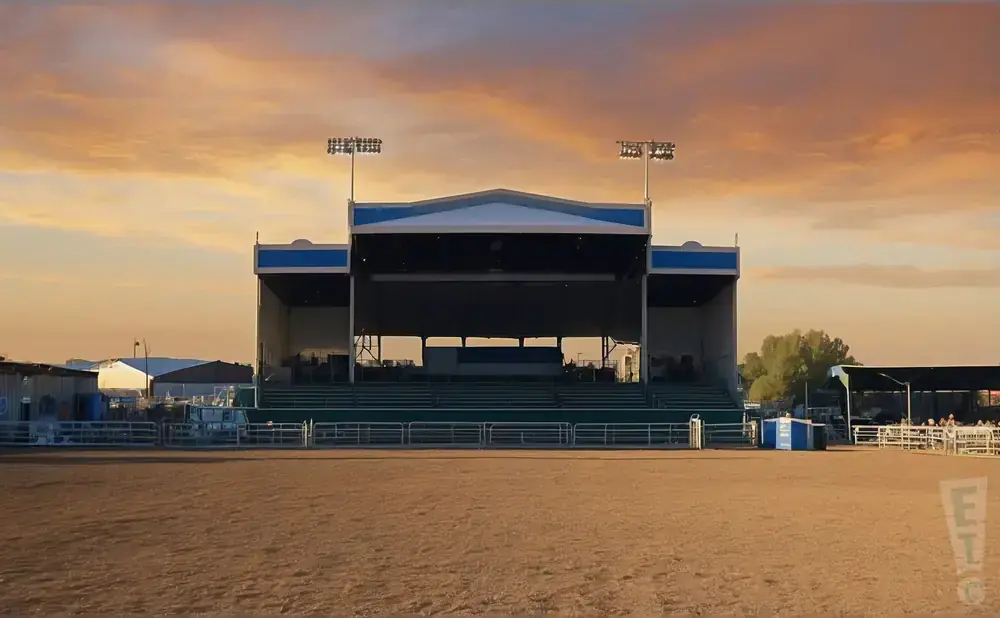 Outdoor amphitheater stage with tiered seating, illuminated by stadium lights under a sunset sky.