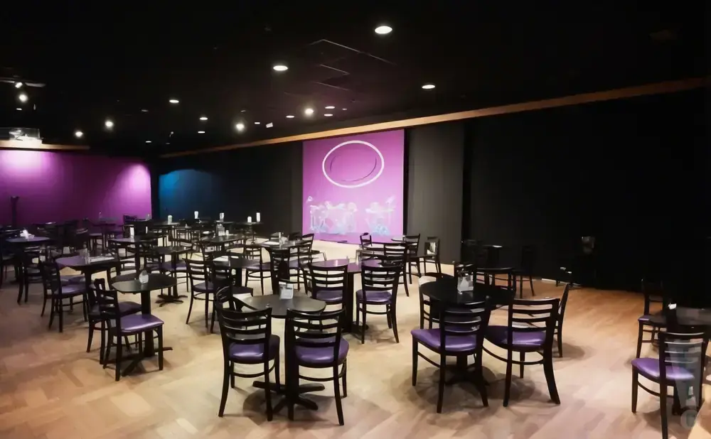 A dimly lit room with many small tables and purple chairs set up for a performance or event.