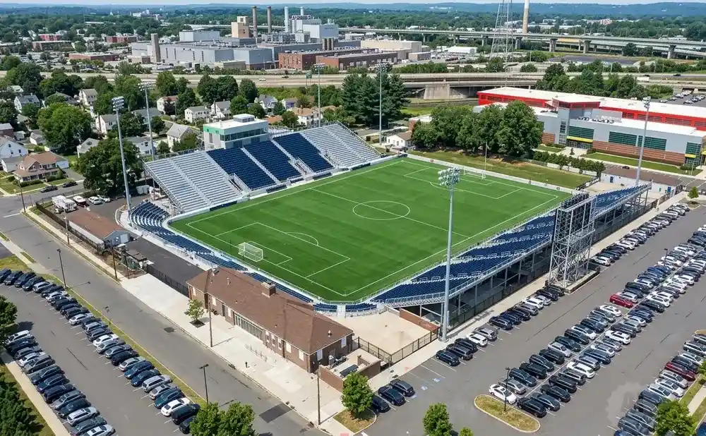 Aerial view of a soccer stadium with a green field, bleacher seating, and surrounding parking lots.