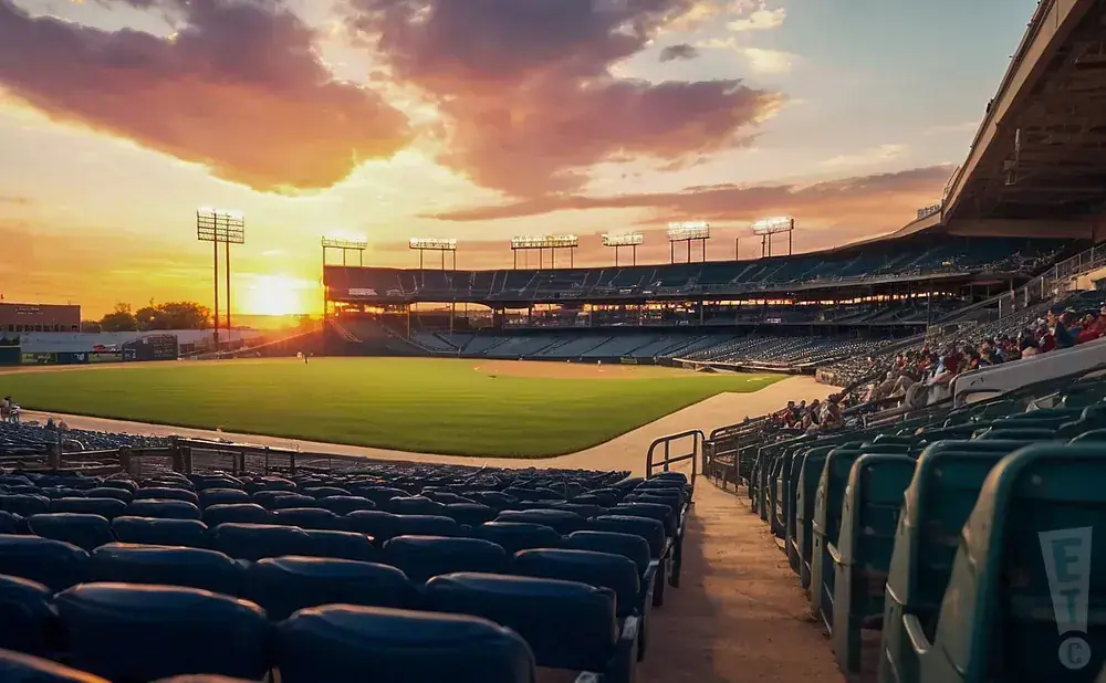 A baseball stadium at sunset, with a vibrant sky over the field and empty seats in the foreground.