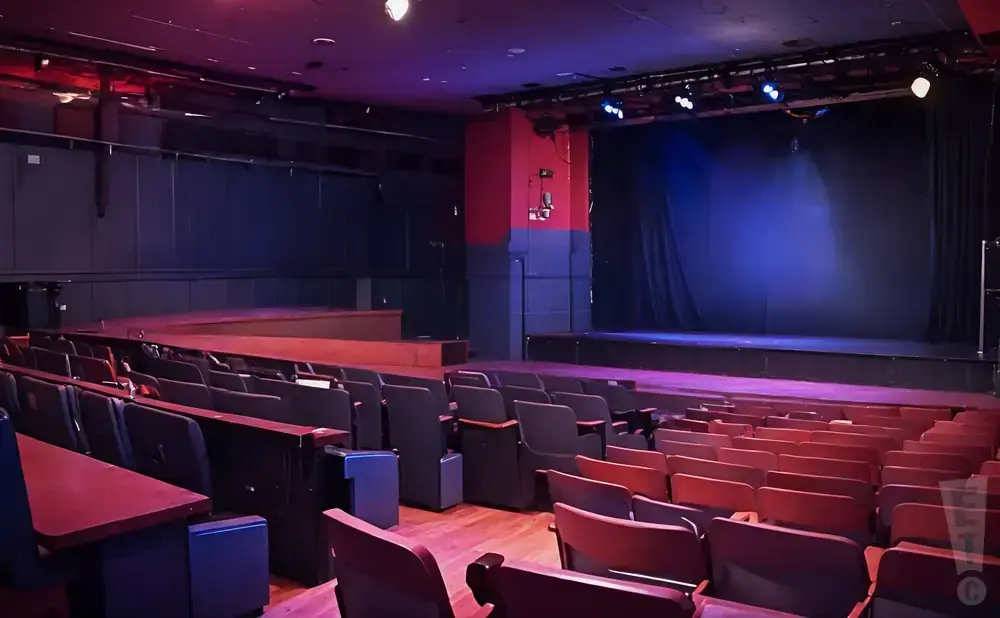 An empty theater with rows of red seats facing a dark stage with blue spotlights.