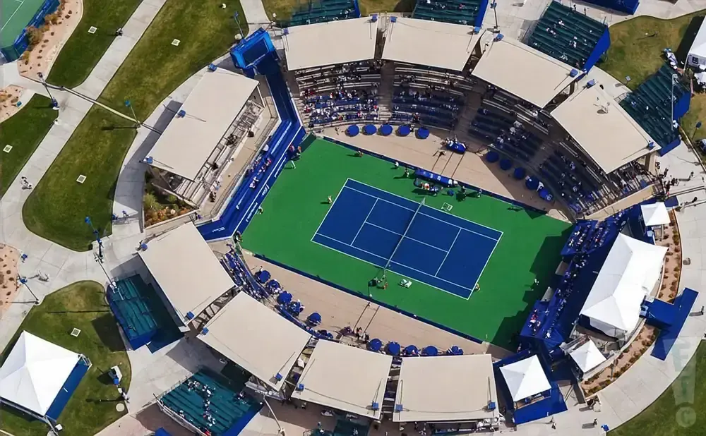 Aerial view of a tennis court with tiered seating and shaded canopies.