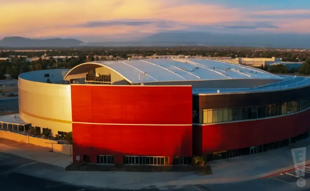 An aerial view of a large, curved, modern building with a white roof and red and tan exterior walls at sunset.