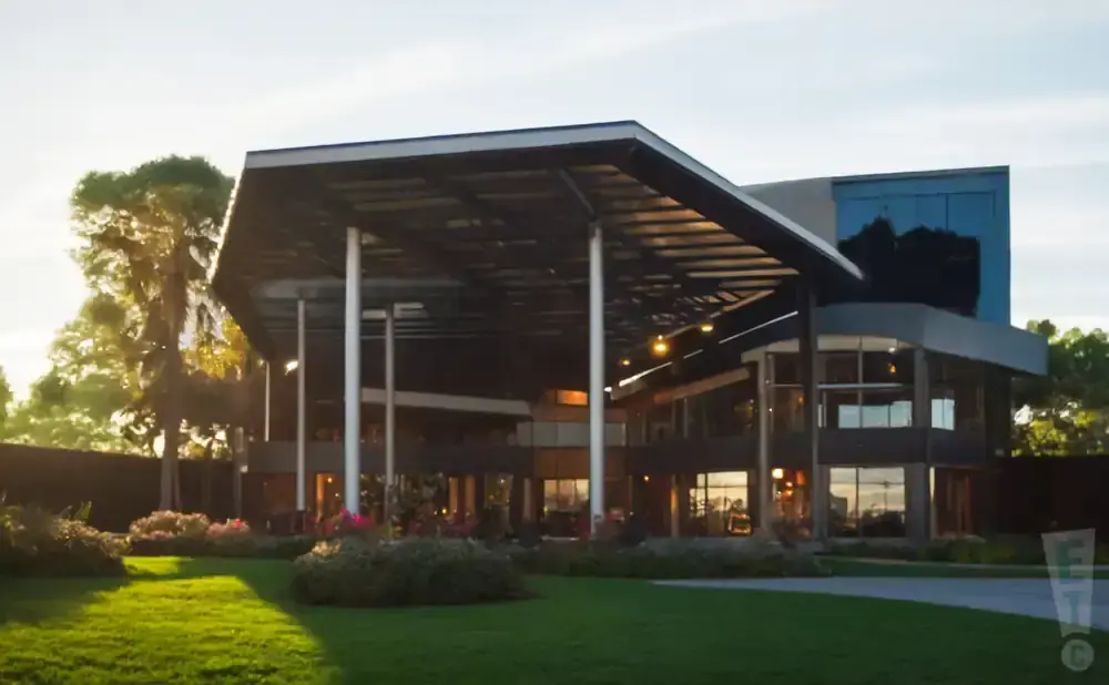 Modern building with large overhang and glass windows, surrounded by lush green lawn and flowering bushes.