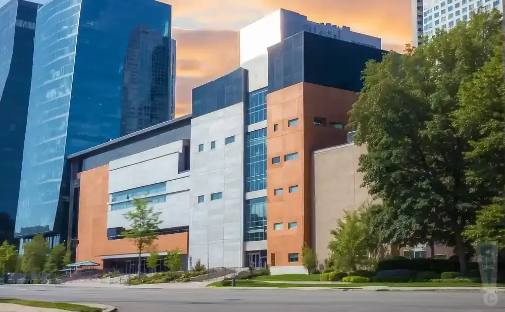 Modern buildings with glass facades and orange accents stand against a sunset sky, with trees in the foreground.