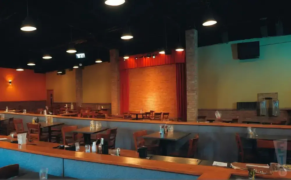 Interior of a restaurant with tables and chairs, a brick stage area with red curtains, and orange and yellow walls.