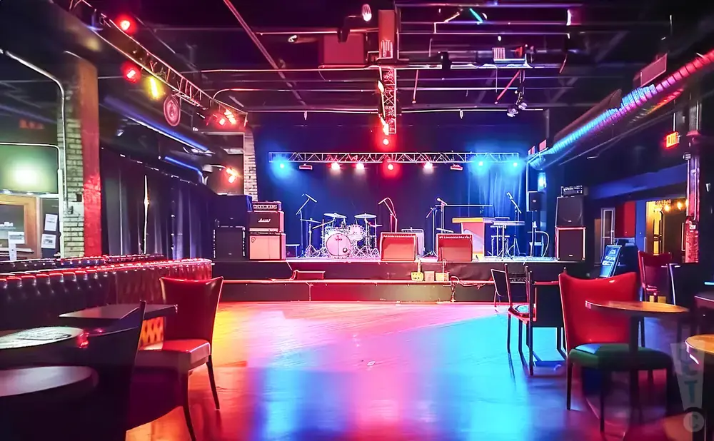 Empty concert venue with stage, drum set, amps, and seating, illuminated by colorful stage lights.