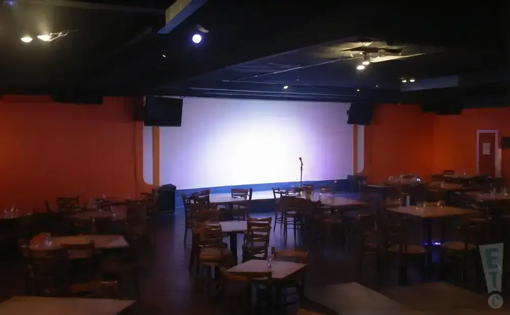 A dimly lit comedy club with orange walls, a large white stage, tables, and chairs.