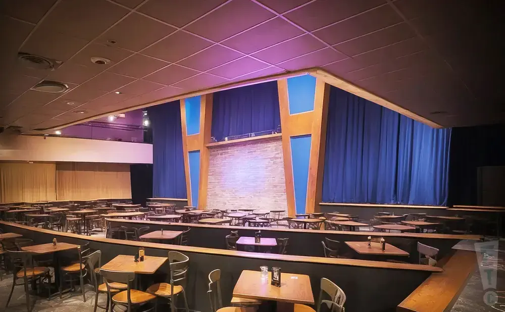 An empty comedy club with tables and chairs facing a stage with blue curtains and a brick backdrop.