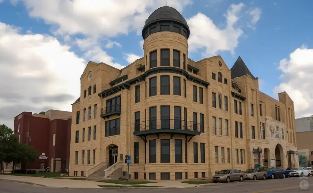 A historic tan brick building with a domed turret and arched entrance stands under a cloudy blue sky.