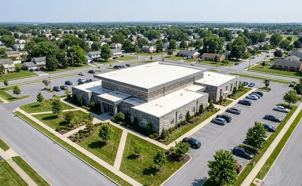 Aerial view of a modern stone-clad building with a flat white roof, surrounded by a parking lot and lush green landscaping, with a suburban neighborhood…