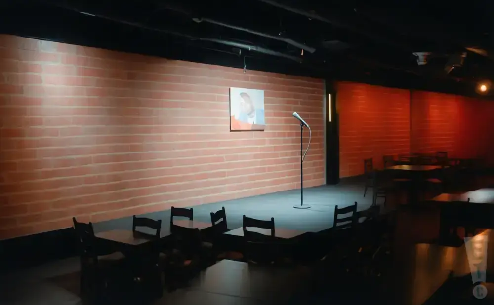 A dimly lit comedy club with a brick wall, microphone, and tables with chairs.