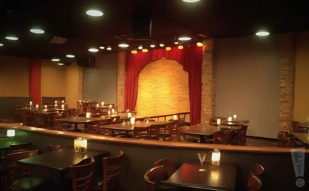 Dark comedy club stage with red curtains and brick backdrop, tables with candles in foreground.