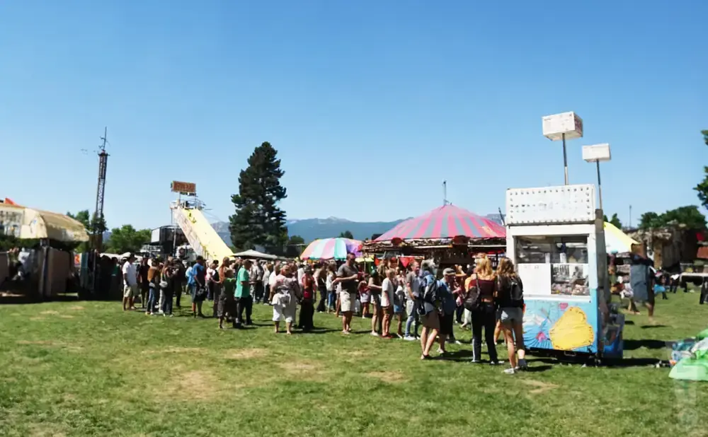 A bustling fairground with a crowd gathered around food stalls and rides under a clear blue sky.