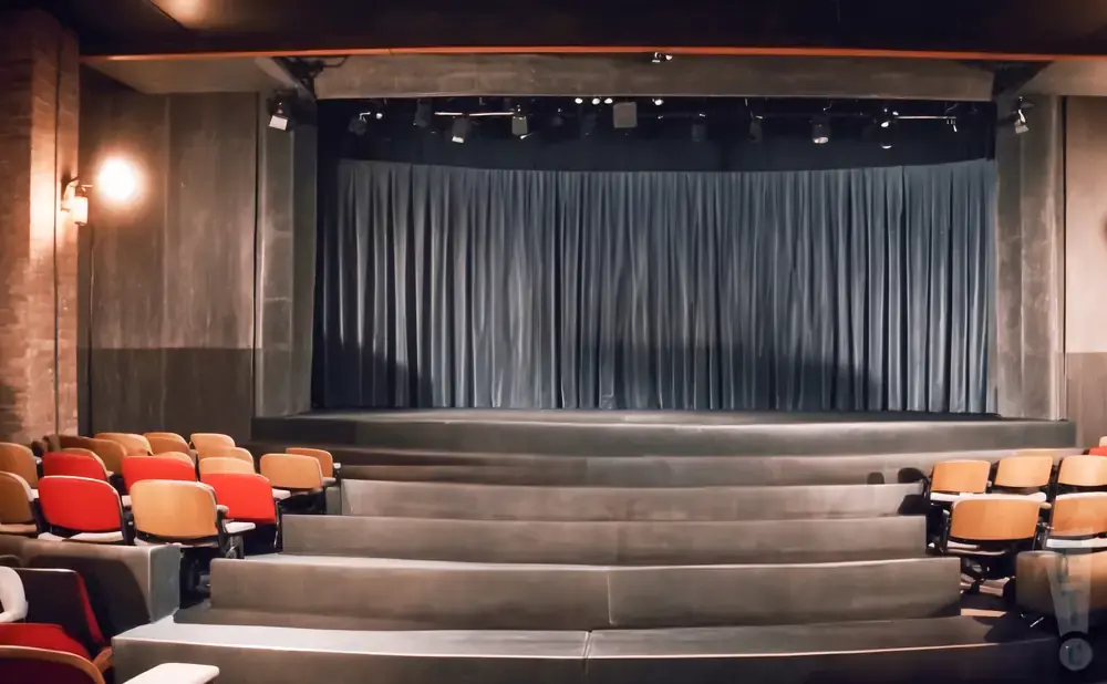 An empty theater with tiered seating facing a stage with blue curtains.