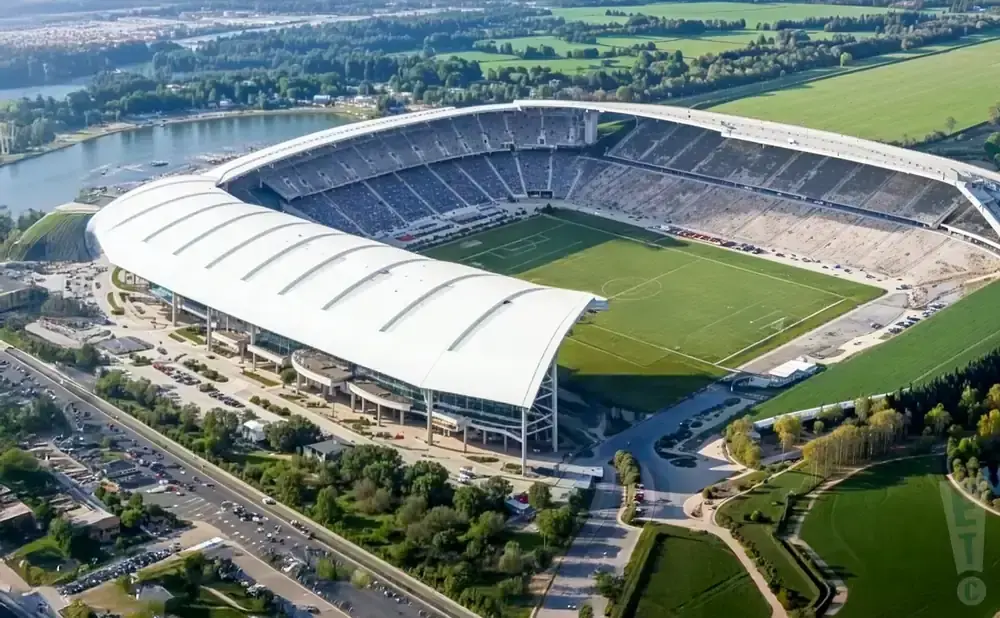 Aerial view of a large stadium with a white roof, surrounded by a football field, parking lots, and a body of water.