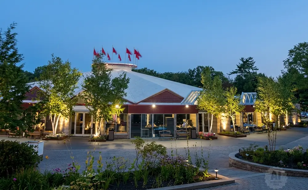Exterior view of a building with a large white roof, red trim, and flags on top, illuminated at dusk.