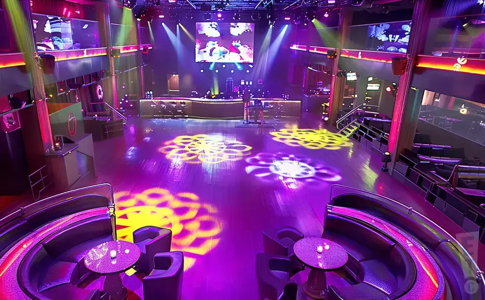 A vibrant nightclub with purple and yellow lights projected onto a wooden floor, and plush seating areas.