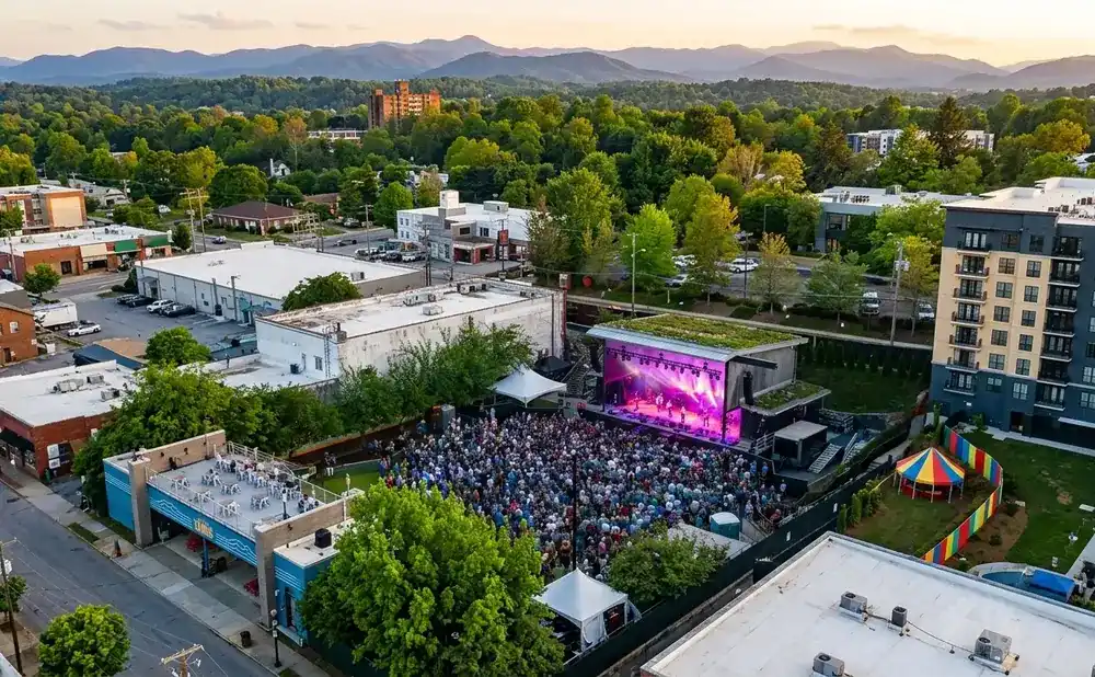 An outdoor concert with a crowd gathered in front of a stage, surrounded by buildings and trees, with mountains in the background.