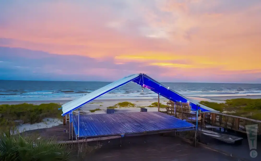 A wooden stage with a blue canopy sits on a beach at sunset.