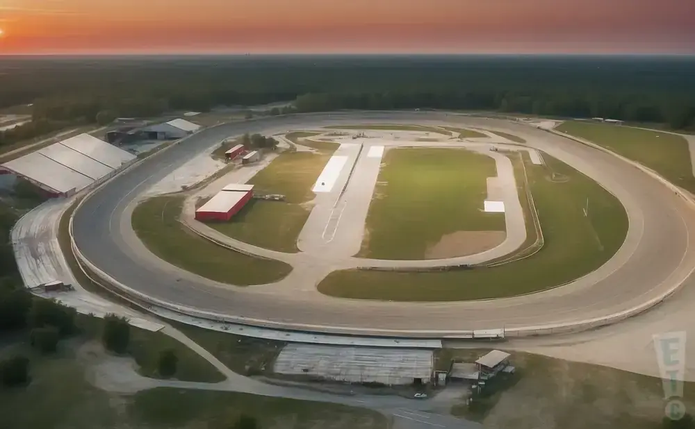 Aerial view of a race track at sunset with a warm orange sky and surrounding trees.
