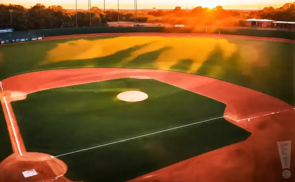 A baseball field at sunset with bright orange light cast across the outfield and a scoreboard in the distance.