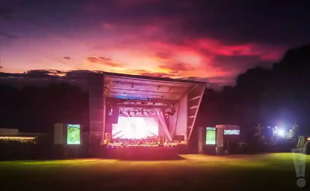 An outdoor stage with a bright screen and lighting is set up on a grassy field, with a vibrant sunset in the background.