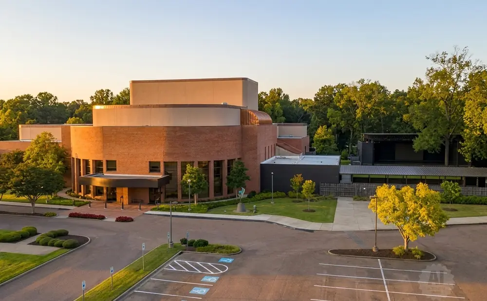 An aerial view of a modern brick building with an outdoor stage area, surrounded by trees during sunset.