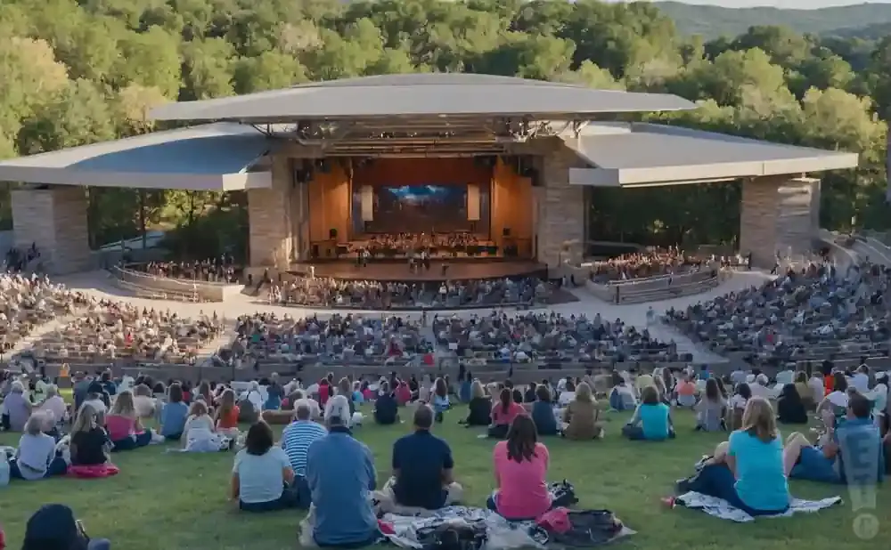 An outdoor amphitheater with a full orchestra performing on stage and a large audience seated in tiered rows and on the grass.