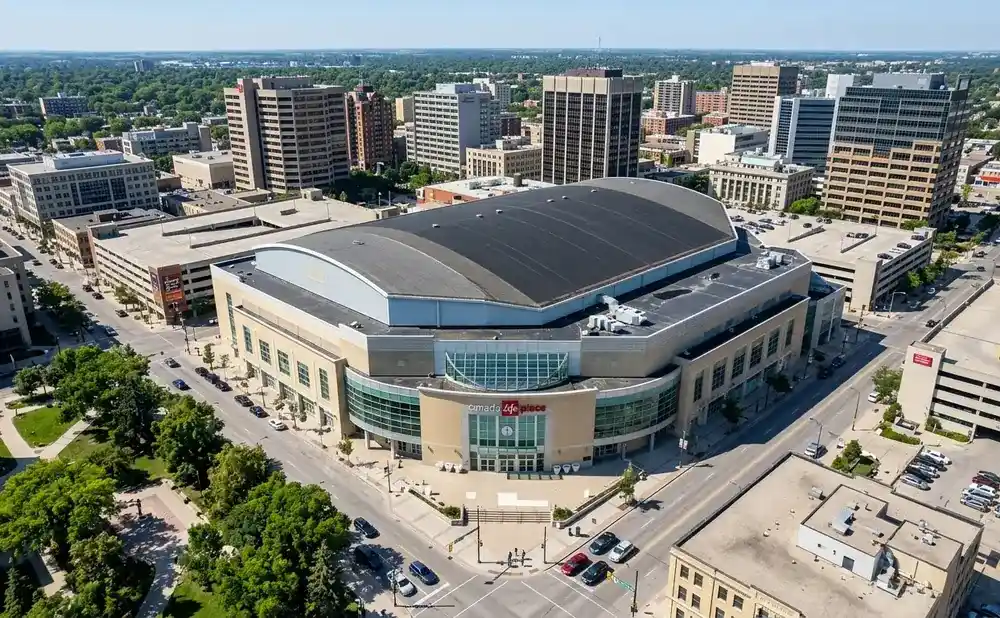 Aerial view of Canada Life Place arena and surrounding downtown buildings under a clear blue sky.