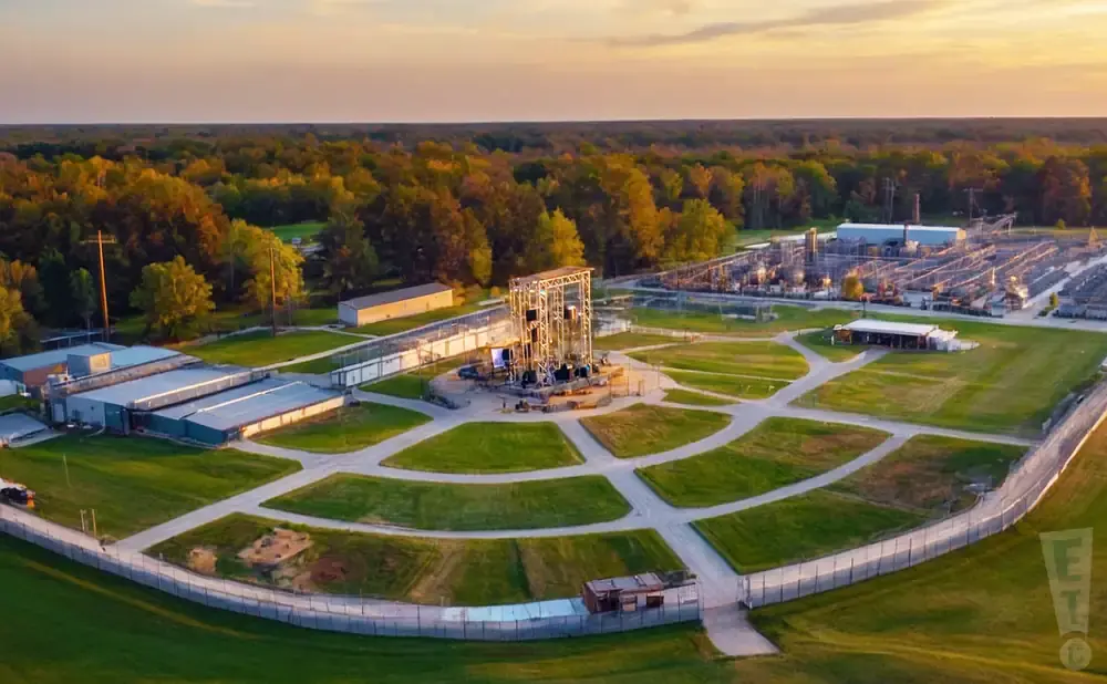Aerial view of a research facility with buildings, a tall metal structure, and surrounding trees at sunset.