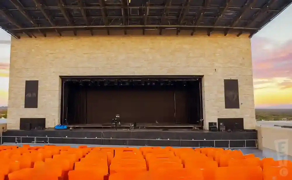 An outdoor amphitheater stage with orange seats in the foreground and a colorful sunset sky.