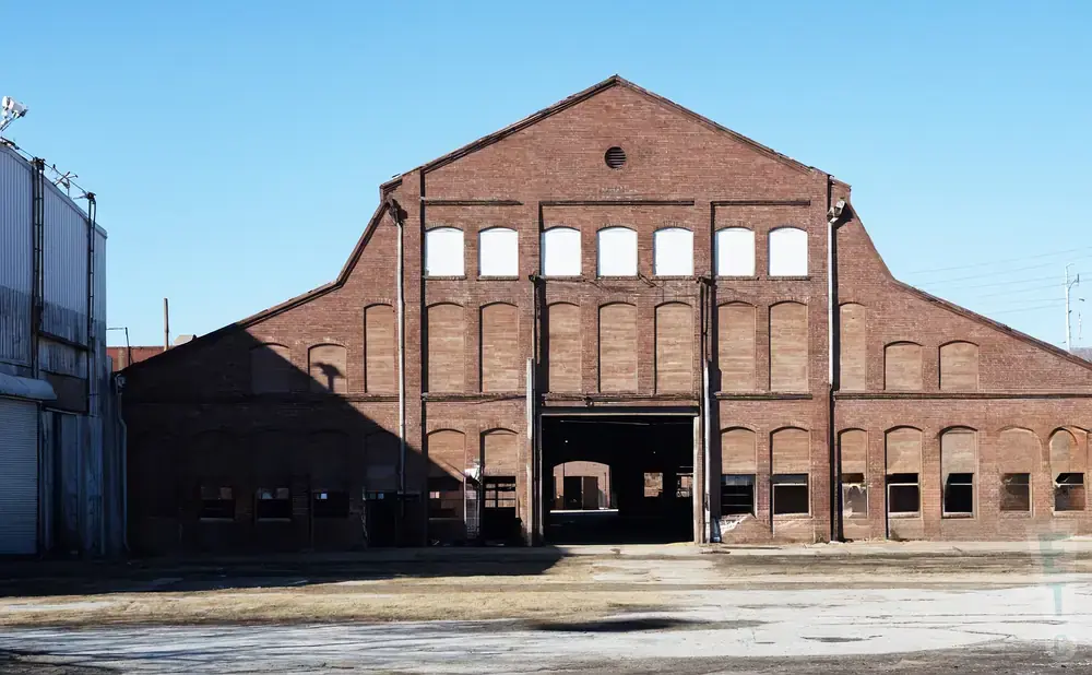 A large, abandoned brick industrial building with a peaked roof and many boarded-up windows.