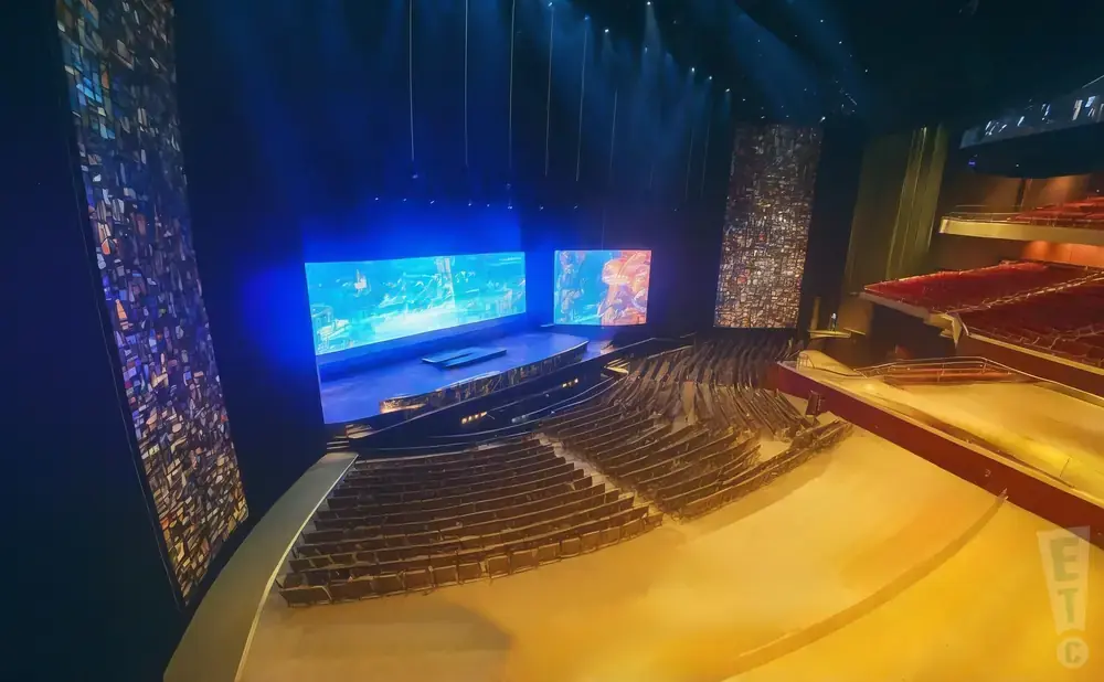An empty theater with a stage, two large screens, and tiered seating.