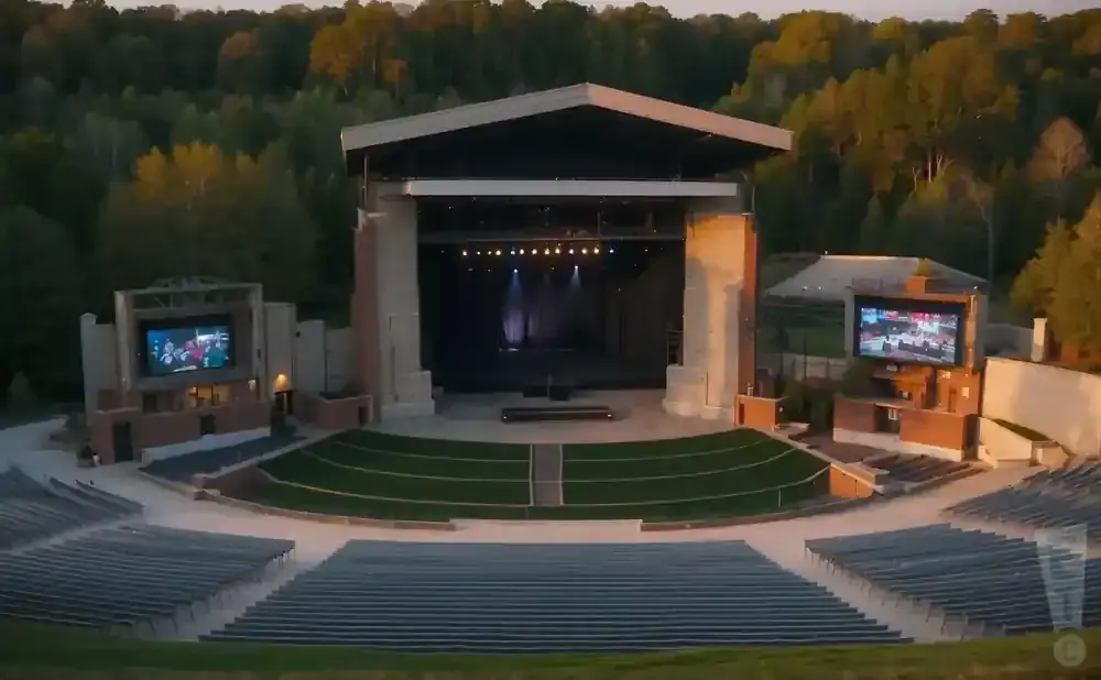 Outdoor amphitheater with a stage, seating, and large screens, surrounded by trees.