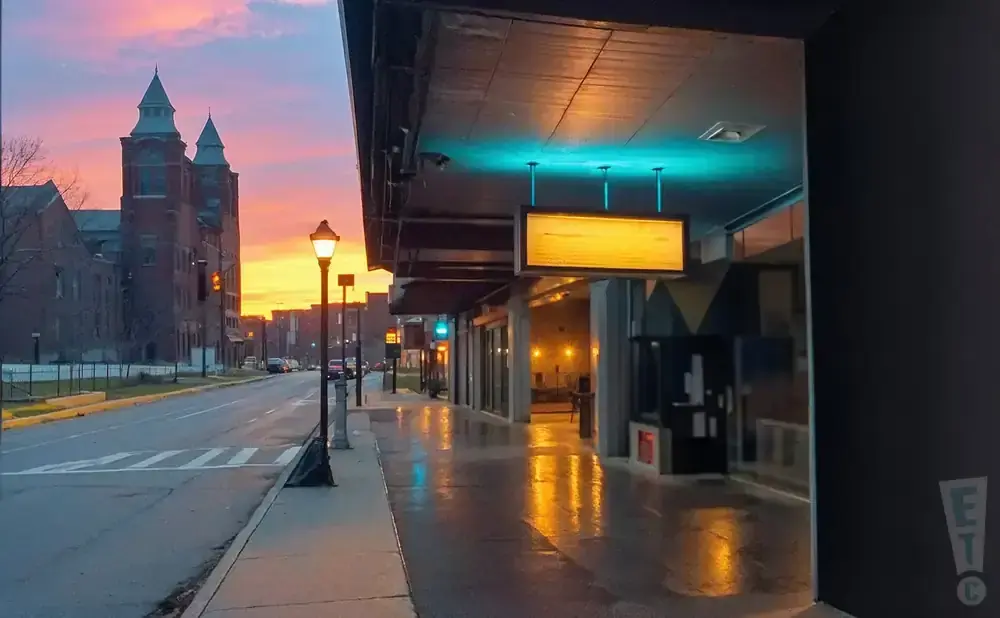 Sunset over a city street with brick buildings, a glowing sign, and wet sidewalks reflecting lights.