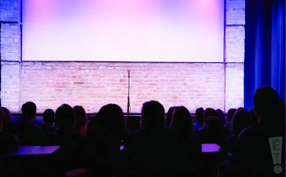 A comedy club stage with a brick wall backdrop, microphone, and audience silhouettes in purple light.
