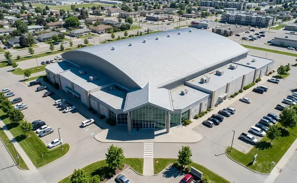 Aerial view of a large arched building with a silver roof, surrounded by parking lots and trees.